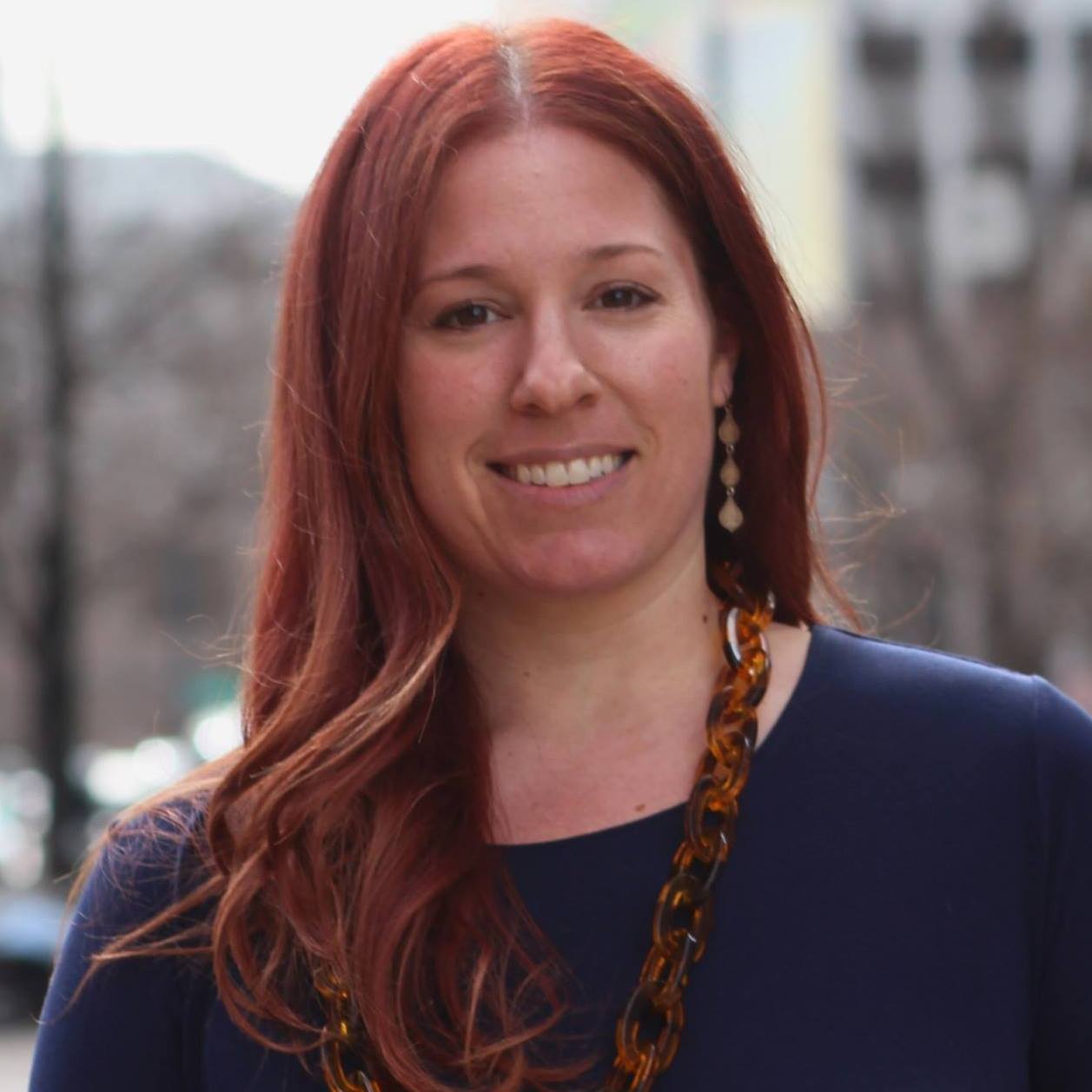 Headshot of Jessica Gronich. A white woman standing in front of the city with red hair.