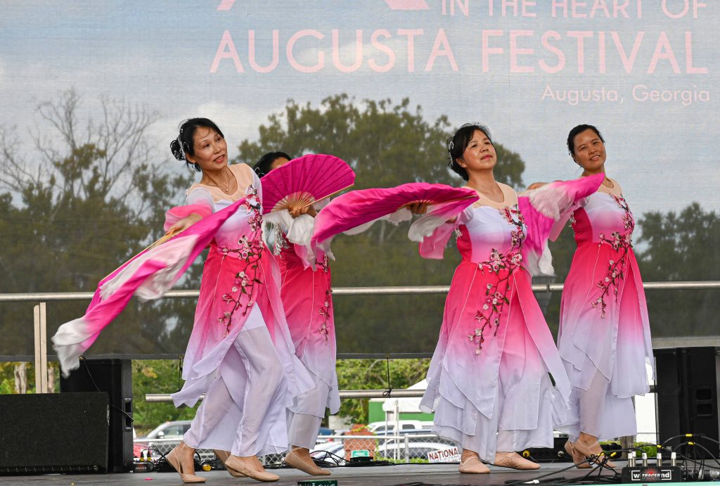 Three women in white and bright pink dancing on stage outside.