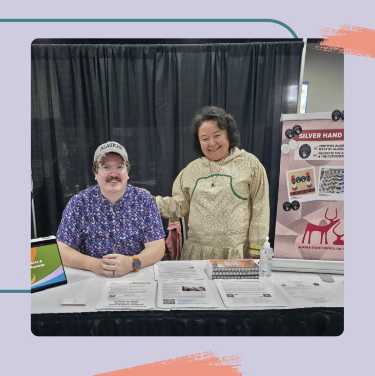 A man sitting at a booth with a blue patterned shirt and gray "ALASKA" cap poses beside a woman in a light traditional outfit as they promote Alaskan arts, featuring a poster that reads "SILVER HAND" behind them.